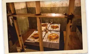 Mesa puesta para el señor cachopo, con platos ámbar y copas, tras unos barrotes.