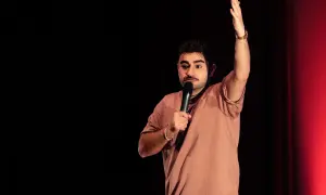 David Puerto Zaragoza, un comediante, levanta la mano mientras sostiene un micrófono en un escenario con cortinas rojas.