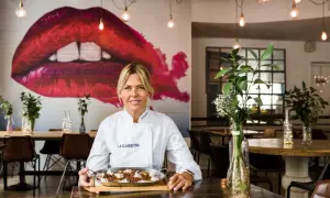 Chef sonriente con paella, perfecta para saber dónde cenar Zaragoza. Mural de labios rojos y luces cálidas crean ambiente.