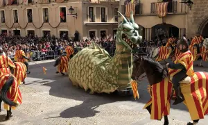 Un dragón gigante desfila en las fiestas de Alcañiz, acompañado por jinetes con trajes de rayas rojas y amarillas.
