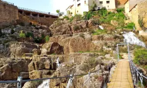 Puente colgante sobre cascadas en Almonacid de la Cuba, con casas tradicionales en la ladera rocosa.