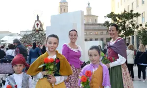 Niñas con trajes regionales y flores rojas, listas para las fiestas Zaragoza. Detrás, un gran tapiz floral y edificios históricos.