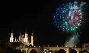 Espectaculares fuegos artificiales iluminan el cielo nocturno sobre el río Ebro, celebrando las fiestas del Pilar. La Basílica del Pilar se alza majestuosa.