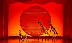 Escena de musicales en Zaragoza: un guepardo y dos jirafas en un escenario con un sol rojo.