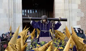 Imagen de una **procesión Miércoles Santo**. Una figura de Jesús carga la cruz, rodeada de nazarenos con túnicas moradas y capirotes dorados.