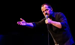 Ángel Martín Zaragoza, comediante, habla al micrófono con gesto expresivo y mano extendida, iluminado por luces azules y púrpuras.