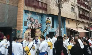 Gente reunida en la calle, algunos con chaquetas blancas y amarillas, celebrando la cincomarzada frente a un edificio con grafitis.