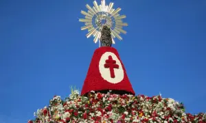 Imagen de una figura religiosa sobre un gran manto de flores rojas y blancas, con un símbolo de cruz. Se celebra una **fiesta** con **pilares** florales.