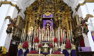 Un altar barroco con imágenes religiosas, velas y flores rojas. Destaca una figura central de Cristo, que parece ser un quinario, rodeado de figuras de la Virgen y otros santos.