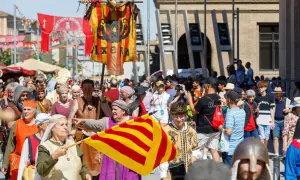 Gente vestida de época desfila en la feria medieval Zaragoza. Un estandarte con un dragón y la palabra "IXERA" ondea.