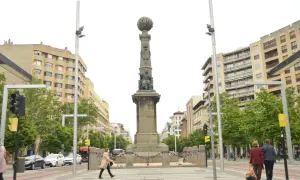 Monumento en la plaza Aragón con edificios modernos y árboles verdes. Dos mujeres caminan con equipaje.
