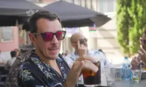 Un hombre con gafas de sol rojas y camisa tropical bebe una bebida en una terraza, quizás disfrutando de un momento de pantomima full Zaragoza.