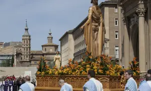 Procesión de **domingo de resurrección Zaragoza**. Figuras religiosas adornadas con flores en un paso, ante edificios históricos y una multitud.