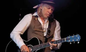 Elliott Murphy Zaragoza, con sombrero y chaleco, toca la guitarra acústica en un concierto.