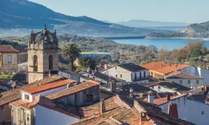 Vista de tejados y un campanario en las Hurdes, con un embalse y montañas al fondo.