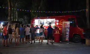 Gente disfrutando en un food trucks festival bajo luces de colores. Un camión rojo sirve comida mientras clientes esperan.