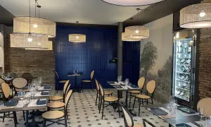 Interior de la brasserie 1885, con mesas puestas, sillas de mimbre y lámparas de ratán. Pared azul y mural de paisaje.