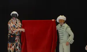 Dos actores con máscaras y trajes de época, uno con un traje de rombos y otro con un traje verde pálido, presentan una tela roja. Parecen parte de una obra de teatro de **els joglars zaragoza**.