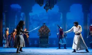 Escena del musical "El Médico" en Zaragoza. Dos hombres se enfrentan con espadas ante un trono dorado, rodeados por otras figuras y un fondo azul.