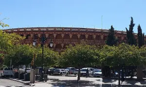 La plaza de toros de Zaragoza, con su arquitectura de arcos y balcones, se alza tras una hilera de árboles frondosos y un aparcamiento con varios coches.