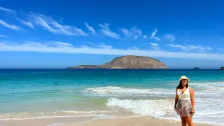 Mujer sonriente en playa de arena dorada, aguas turquesas y una isla montañosa al fondo. ¡Un día perfecto para ver qué ver en La Graciosa!