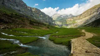 Un sendero de piedra cruza un prado verde junto a un río en un valle montañoso. Un paisaje espectacular que ver en España.