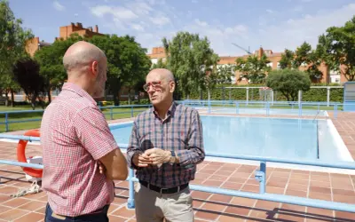 Dos hombres conversan junto a unas piscinas en Zaragoza. El sol brilla en un día despejado.