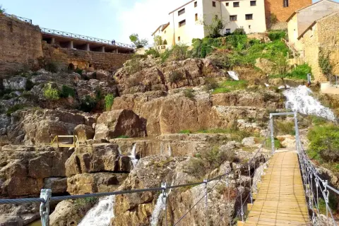 Puente colgante sobre cascadas en Almonacid de la Cuba, con casas tradicionales en la ladera rocosa.