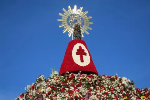 Imagen de una figura religiosa sobre un gran manto de flores rojas y blancas, con un símbolo de cruz. Se celebra una **fiesta** con **pilares** florales.