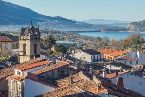 Vista de tejados y un campanario en las Hurdes, con un embalse y montañas al fondo.