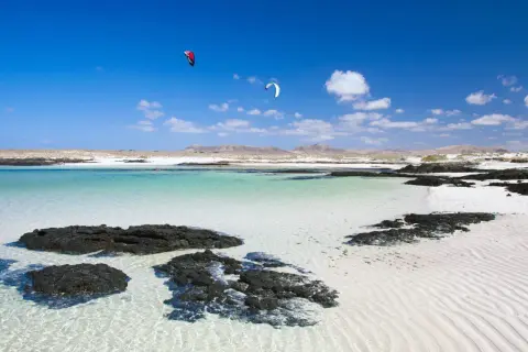 Aguas turquesas y arena blanca en Fuerteventura, con kitesurfistas en el horizonte. Un paraíso entre los 10 sitios para visitar en Fuerteventura.
