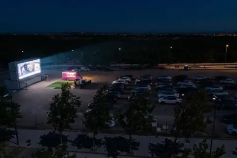 Noche de cine de verano Zaragoza. Coches aparcados frente a una gran pantalla donde proyectan una película, con un camión de comida rosa al lado.