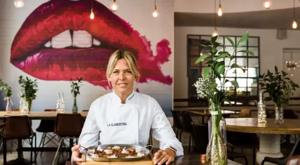 Chef sonriente con paella, perfecta para saber dónde cenar Zaragoza. Mural de labios rojos y luces cálidas crean ambiente.