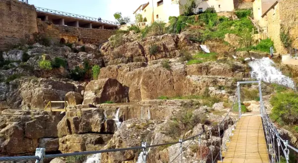Puente colgante sobre cascadas en Almonacid de la Cuba, con casas tradicionales en la ladera rocosa.