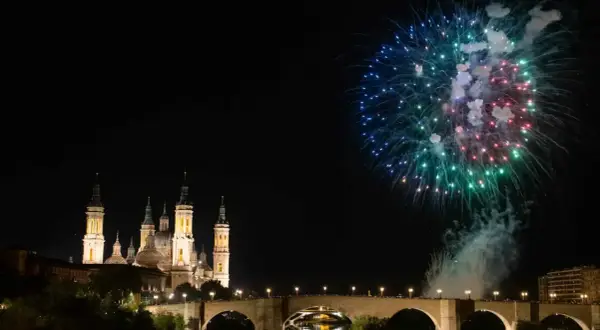 Espectaculares fuegos artificiales iluminan el cielo nocturno sobre el río Ebro, celebrando las fiestas del Pilar. La Basílica del Pilar se alza majestuosa.