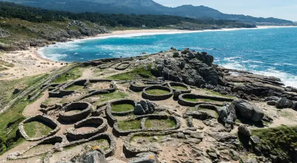 Ruinas circulares de un castro gallego, un lugar histórico que ver en A Coruña, con el mar azul y la costa de fondo.