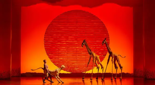 Escena de musicales en Zaragoza: un guepardo y dos jirafas en un escenario con un sol rojo.