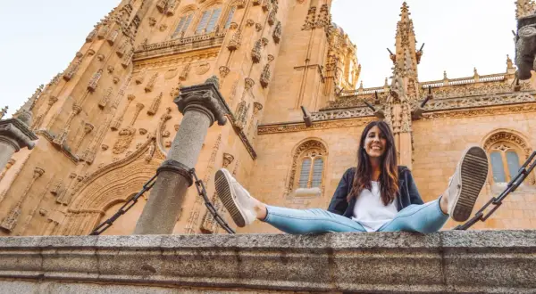 Una mujer sonríe sentada en un muro de piedra frente a la imponente fachada de la Catedral de Salamanca, un lugar clave que ver en Salamanca.