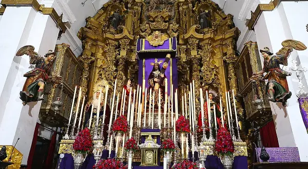 Un altar barroco con imágenes religiosas, velas y flores rojas. Destaca una figura central de Cristo, que parece ser un quinario, rodeado de figuras de la Virgen y otros santos.