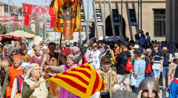 Gente vestida de época desfila en la feria medieval Zaragoza. Un estandarte con un dragón y la palabra "IXERA" ondea.