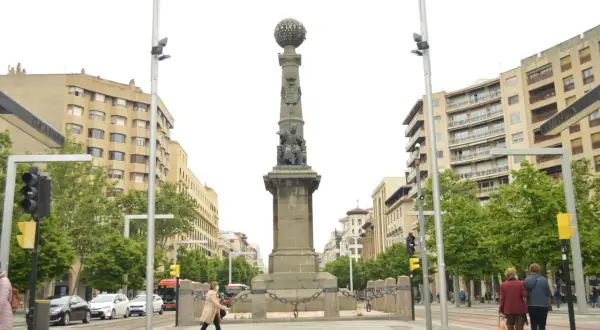 Monumento en la plaza Aragón con edificios modernos y árboles verdes. Dos mujeres caminan con equipaje.