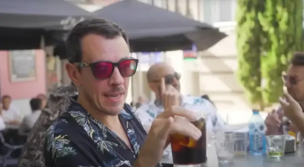 Un hombre con gafas de sol rojas y camisa tropical bebe una bebida en una terraza, quizás disfrutando de un momento de pantomima full Zaragoza.