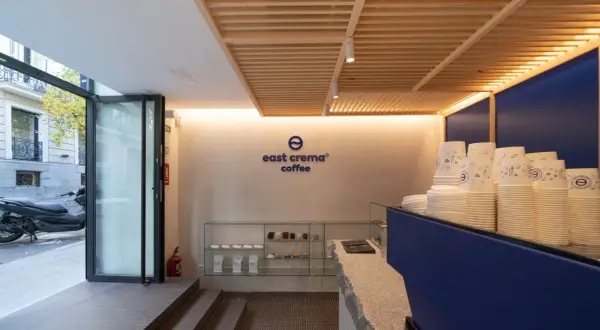 Interior de cafetería con logo "east crema coffee", vasos apilados y estanterías con productos.