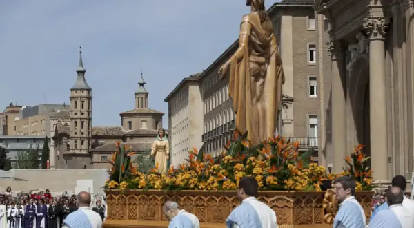 Procesión de **domingo de resurrección Zaragoza**. Figuras religiosas adornadas con flores en un paso, ante edificios históricos y una multitud.