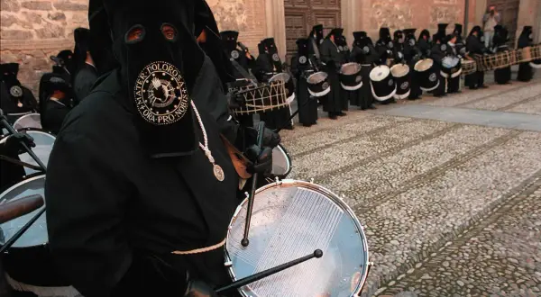 Tambores resuenan en las procesiones de Zaragoza. Nazarenos con túnicas negras y capirotes golpean sus tambores, creando un ritmo solemne.
