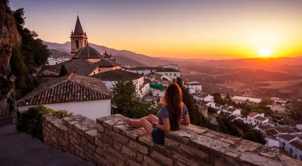 Una mujer contempla el atardecer sobre un pueblo blanco, con tejados de los mayses y montañas al fondo.