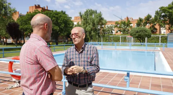 Dos hombres conversan junto a unas piscinas en Zaragoza. El sol brilla en un día despejado.