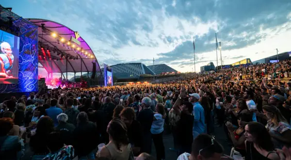 Multitud disfrutando de un concierto al aire libre. Se ve un gran escenario con luces y pantallas, y miles de personas animando. ¡Así se vive latino Zaragoza!