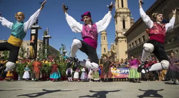 Jóvenes bailan jotas del Pilar con castañuelas, saltando enérgicamente. Al fondo, gente vestida de época y edificios históricos.
