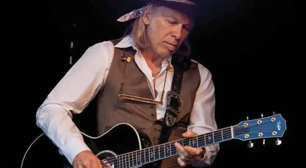 Elliott Murphy Zaragoza, con sombrero y chaleco, toca la guitarra acústica en un concierto.